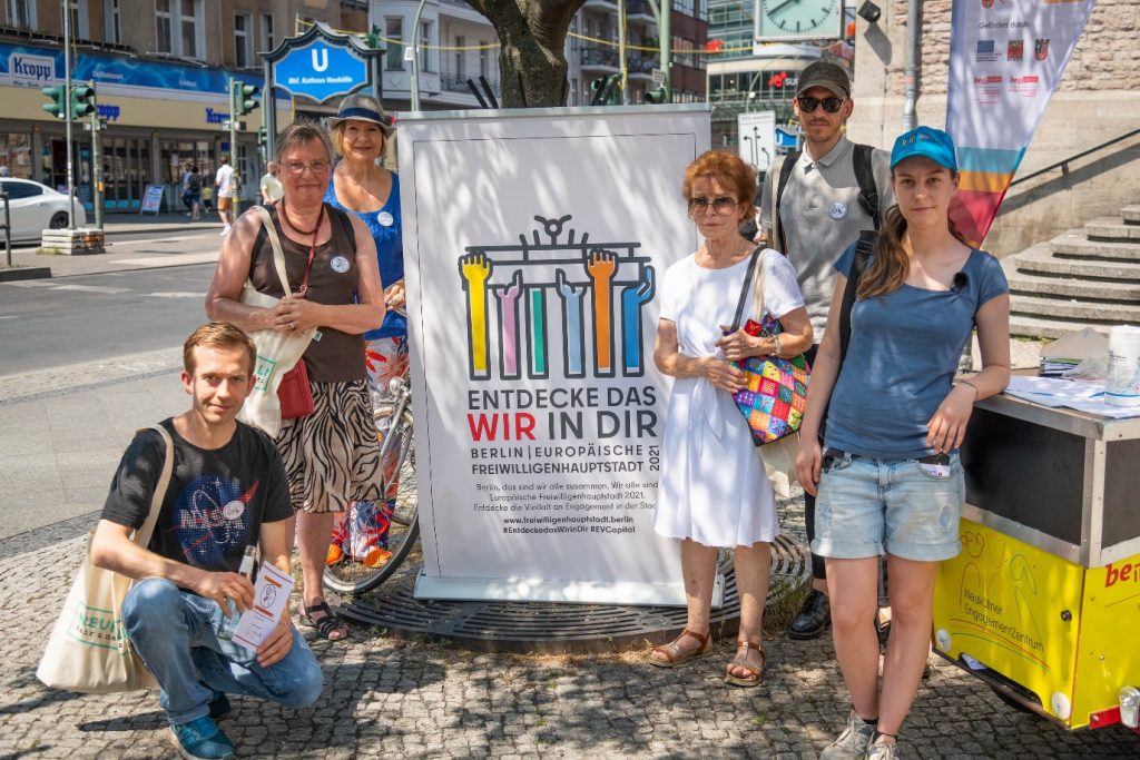 Das Bild zeigt eine Gruppe von Rallye-Teilnehmenden vor einem Roll-Up mit dem Schriftzug "Entdecke das wir in dir - Berlin Europäische Freiwilligenhauptstadt 2021". 